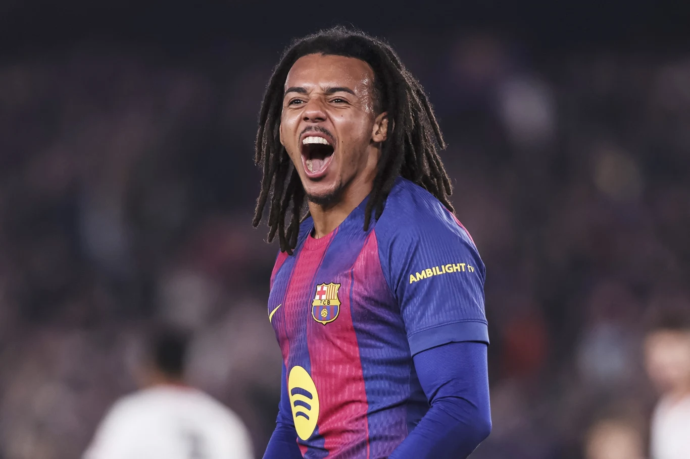 Piłkarz w stroju Barcelony z rozpostartymi ramionami i wyrażającą silne emocje twarzą, na tle rozmazanej publiczności stadium.