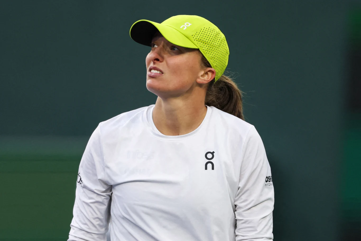 Kobieta ubrana w sportową odzież oraz jasną czapkę z daszkiem patrząca w górę, z zielonym tłem rozmytym na drugim planie, podkreślającym postać.