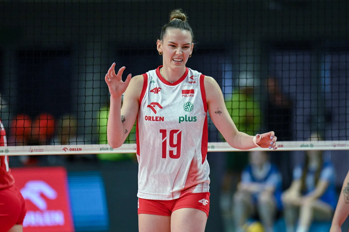 Zawodniczka polskiej reprezentacji siatkówki ubrana w biało-czerwony strój sportowy z numerem 19, stoi przy siatce na hali sportowej, unosząc ręce w geście reakcji podczas meczu.