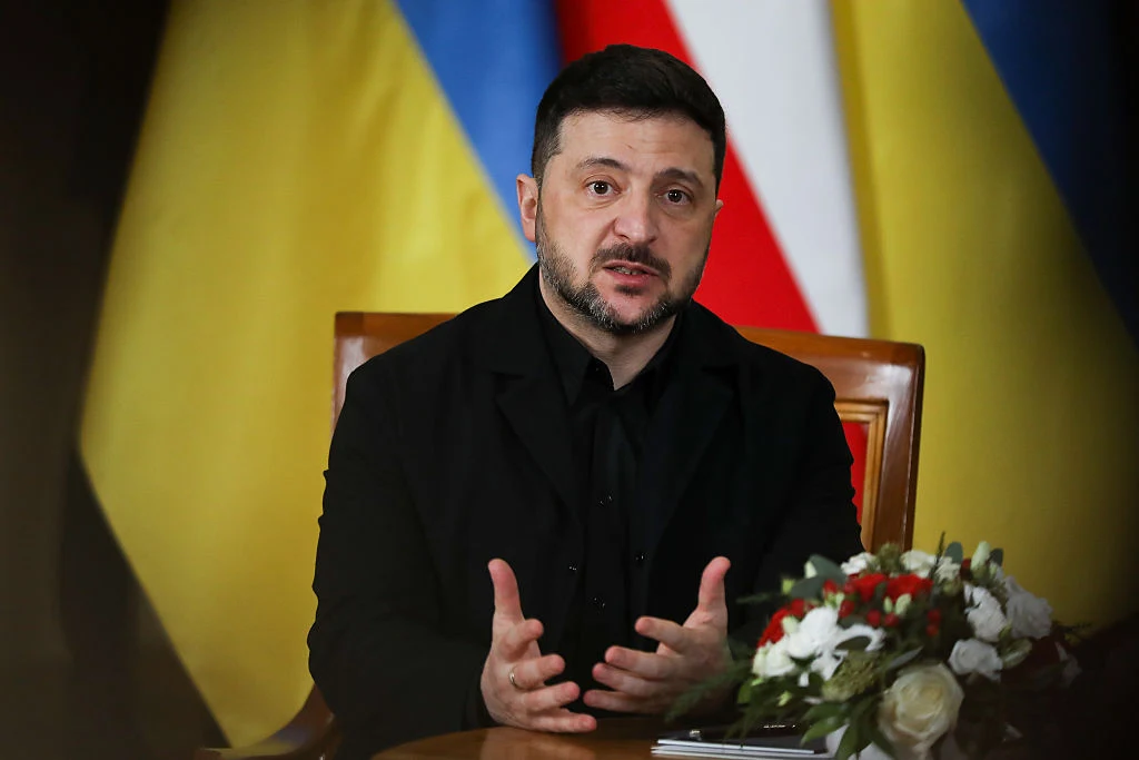 Wojna w Ukrainie. Wołodymyr Zełenski zapowiada kompromisy Mężczyzna w ciemnym garniturze siedzi przy stole, gestykulując podczas rozmowy. W tle widać flagi Ukrainy i Polski. Na stole stoi bukiet kwiatów oraz dokumenty.