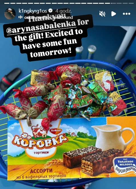 Słodycze Kyrgiosa Kolorowe cukierki oraz pudełko wafelków marki Korowka umieszczone na niebieskiej rakiecie tenisowej, w tle fragmenty boiska tenisowego.