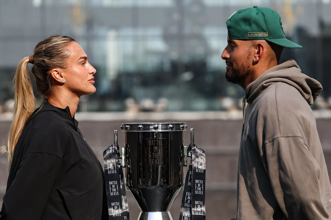 Aryna Sabalenka, Nick Kyrgios Dwie osoby stoją naprzeciw siebie i patrzą sobie w oczy, między nimi ustawiony jest duży, srebrny puchar na tle nowoczesnego budynku.
