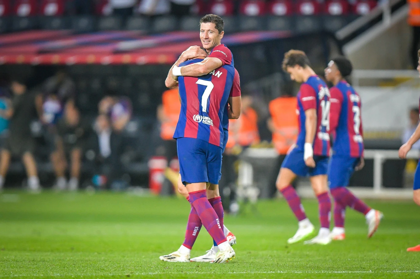 Piłkarze w bordowo-granatowych strojach świętują zdobycie bramki na stadionie, jeden z nich obejmuje drugiego, a w tle widoczni są pozostali zawodnicy i kibice na trybunach.