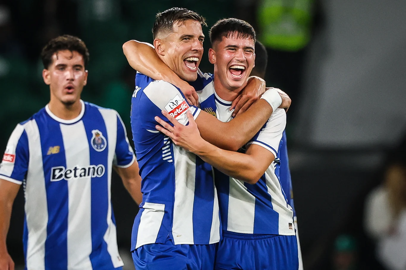 Dwóch piłkarzy w niebiesko-białych strojach ściska się i uśmiecha z radości po udanym momencie w meczu, w tle widoczny jeszcze jeden zawodnik oraz rozmazane postaci na trybunach.