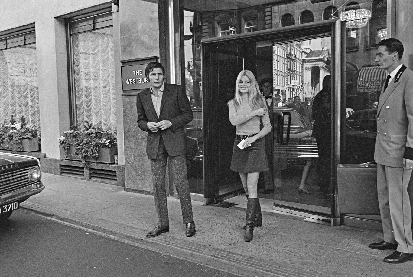 Przed wejściem do budynku hotelowego stoi elegancka kobieta w krótkiej sukience i wysokich butach, obok niej mężczyzna ubrany w marynarkę i spodnie, a po prawej stronie widoczny jest portier w uniformie. Z wnętrza hotelu pada światło na ulicę, na które...