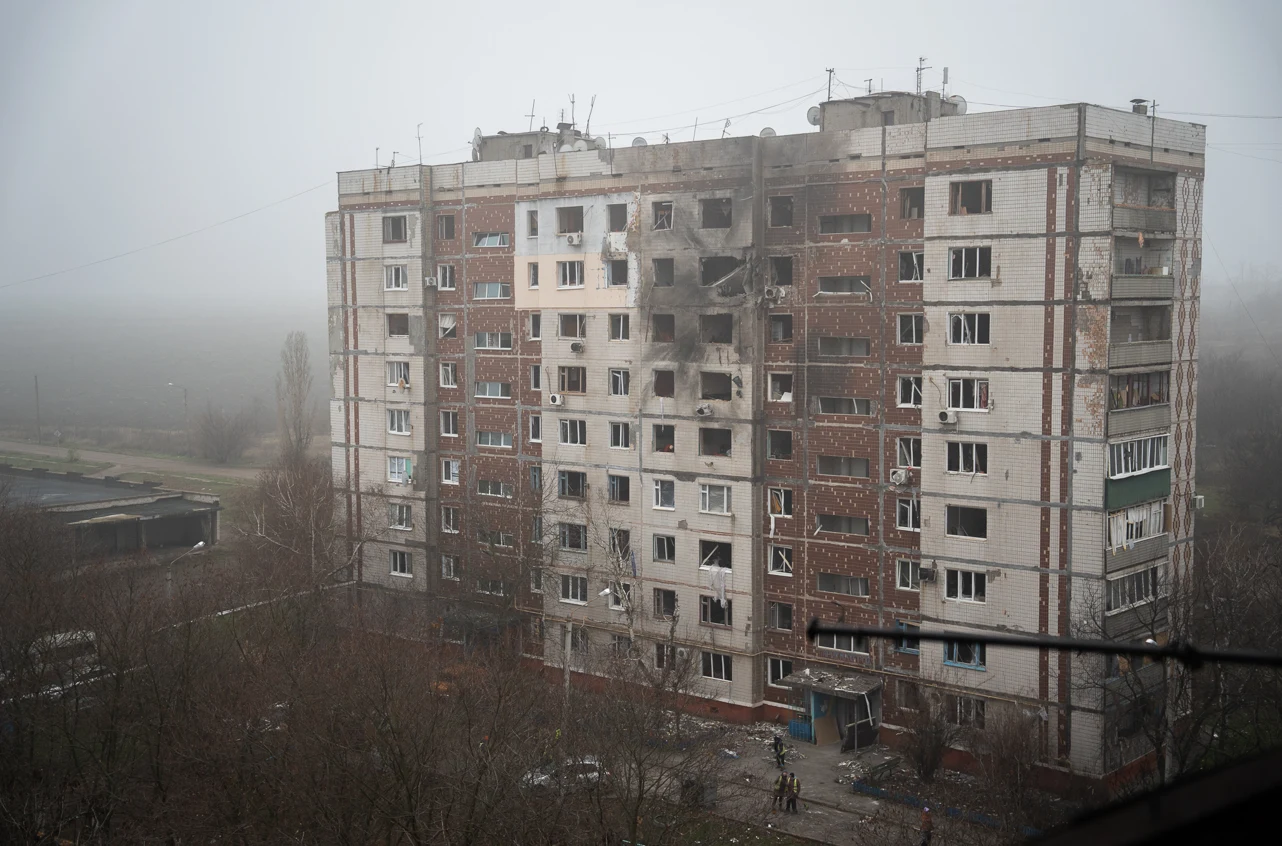Wielopiętrowy budynek mieszkalny o zniszczonej elewacji, z licznymi śladami po eksplozjach lub ostrzałach, na tle zamglonego, szarego krajobrazu. Wokół budynku niewielka liczba drzew bez liści i widoczna pusta przestrzeń.