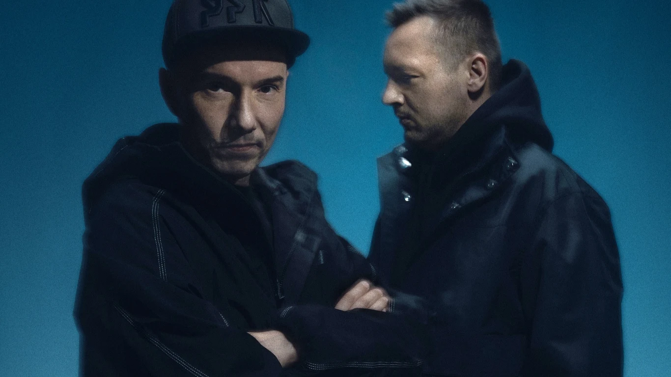 Rahim i Fokus Dwóch mężczyzn ubranych w ciemne bluzy i kurtki, jeden patrzy wprost, drugi bokiem, tło utrzymane w niebieskiej tonacji.