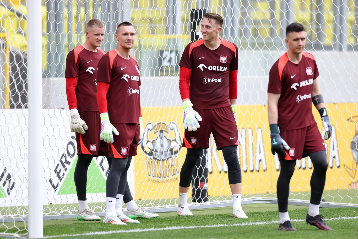 Bartosz Mrozek, Łukasz Skorupski, Marcin Bułka i Mateusz Kochalski na treningu reprezentacji Polski Czterech bramkarzy ubranych w bordowe stroje treningowe reprezentacji Polski stoi w bramce na stadionie piłkarskim, rozmawia i przygotowuje się do treningu, w tle widoczne siatka oraz fragmenty trybun.