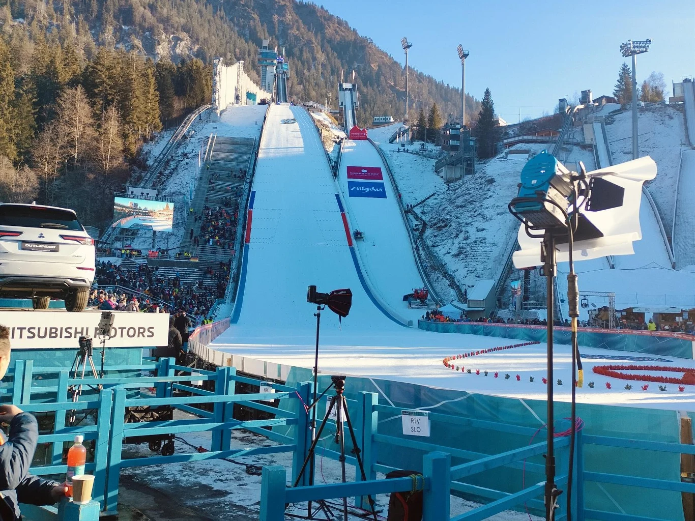 Skocznia narciarska otoczona górskim krajobrazem, z ustawioną aparaturą fotograficzną oraz samochodem na platformie promocyjnej. Liczni kibice zgromadzeni wokół obiektu, trybuny wypełnione. Oświetlenie i telebimy dodają atmosfery sportowej imprezy.