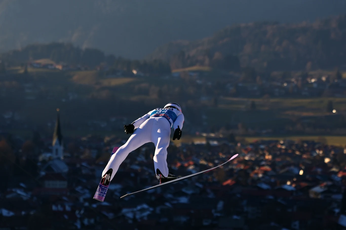 Paweł Wąsek Skoczek narciarski w locie nad miastem, z rozległym widokiem na dolinę i góry w tle, w pełnym stroju sportowym i numerze startowym, dynamiczny moment w czasie zawodów.