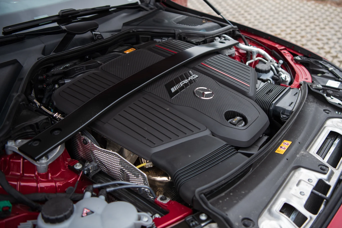 Silnik samochodu marki Mercedes-AMG z widocznym logo producenta, czarną obudową i elementami konstrukcyjnymi, otoczony częściami karoserii w kolorze bordowym.