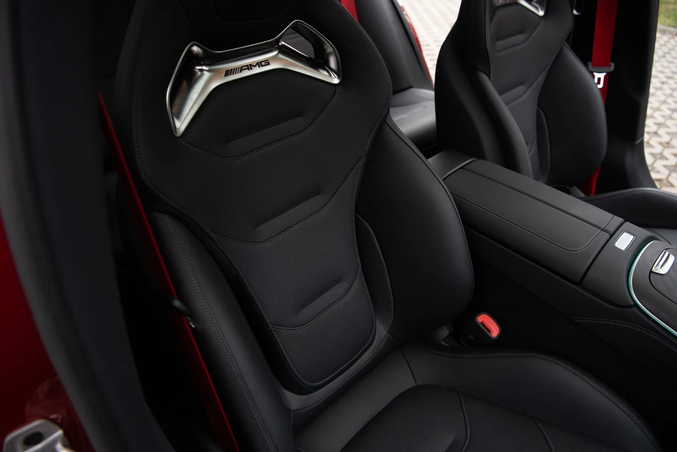 Czarne sportowe fotele samochodowe z logo AMG, wykończone wysokiej jakości materiałem, z metalowymi wstawkami na zagłówkach, umieszczone we wnętrzu auta z czerwonymi akcentami na drzwiach.