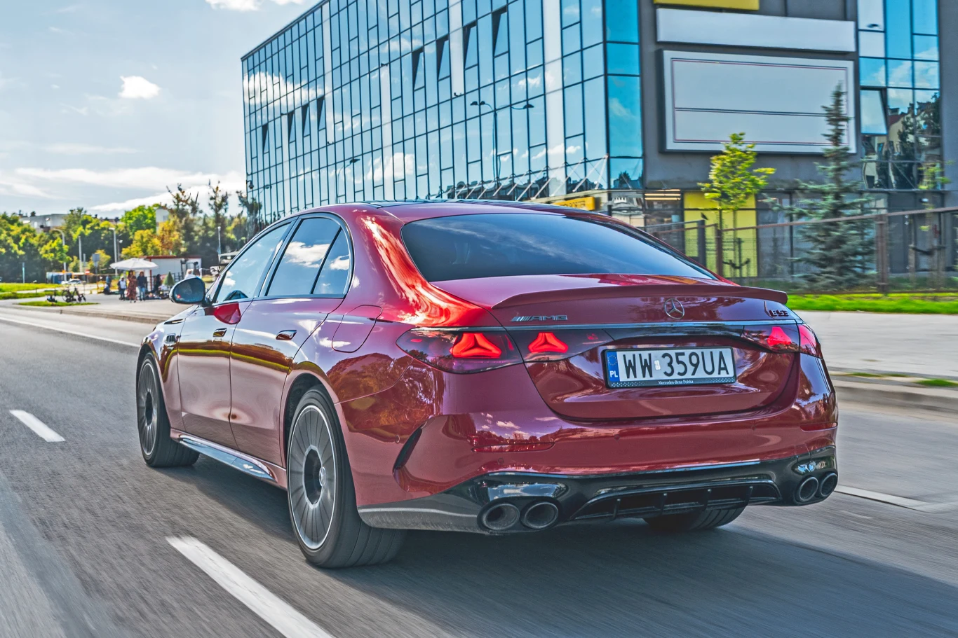 Czerwony luksusowy sedan jadący po miejskiej ulicy, w tle widoczny nowoczesny szklany biurowiec oraz rozmyte drzewa i chodnik.