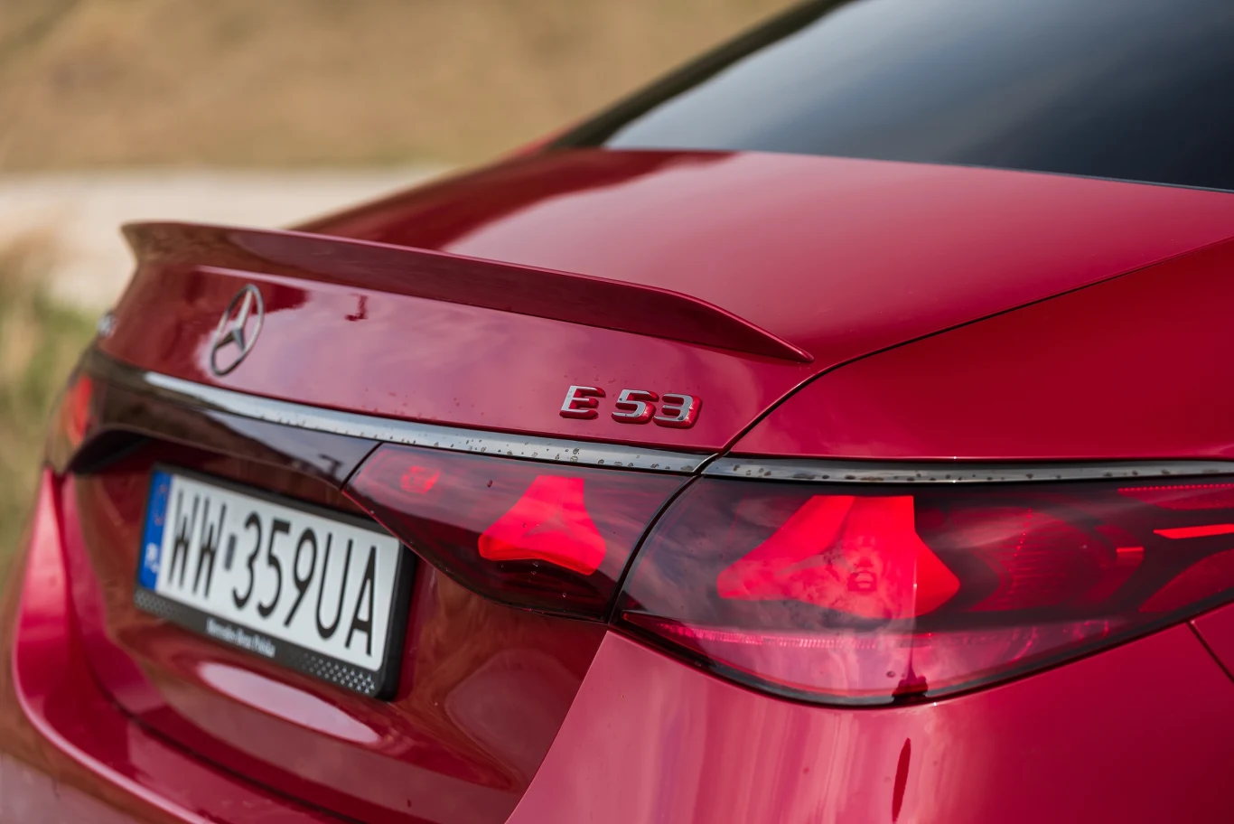 Tylna część czerwonego samochodu marki Mercedes-Benz model E 53 AMG z widocznym logo, emblematem modelu oraz świecącymi światłami tylnymi, na tle rozmytego krajobrazu, z tablicą rejestracyjną oznaczoną napisem ‚UA’.