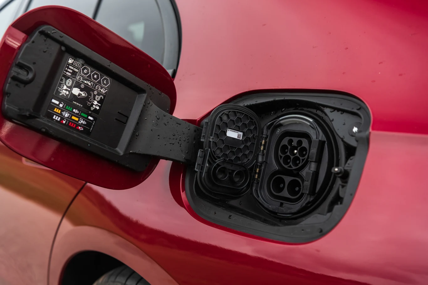 Port ładowania samochodu elektrycznego wraz z naklejką informacyjną na klapce, na tle czerwonej karoserii pojazdu. Złącze wyposażone w dwa różne wejścia do ładowania oraz widoczne oznaczenia techniczne.