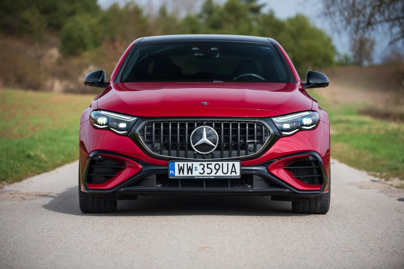 Czerwony samochód marki Mercedes z widocznym przednim grillem oraz charakterystycznym logo, ustawiony na wiejskiej drodze, otoczony zielenią, z czarnymi detalami nadwozia i nowoczesnym wyglądem reflektorów.