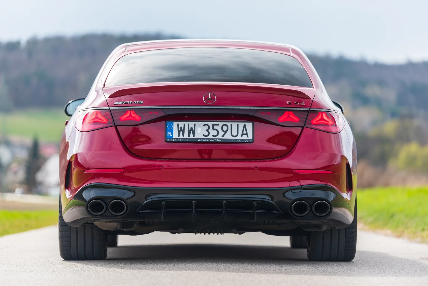 Czerwony samochód marki Mercedes z widocznym napisem AMG i tablicą rejestracyjną WW 359UA, sfotografowany od tyłu na wiejskiej drodze z nieostrym tłem naturalnego krajobrazu.