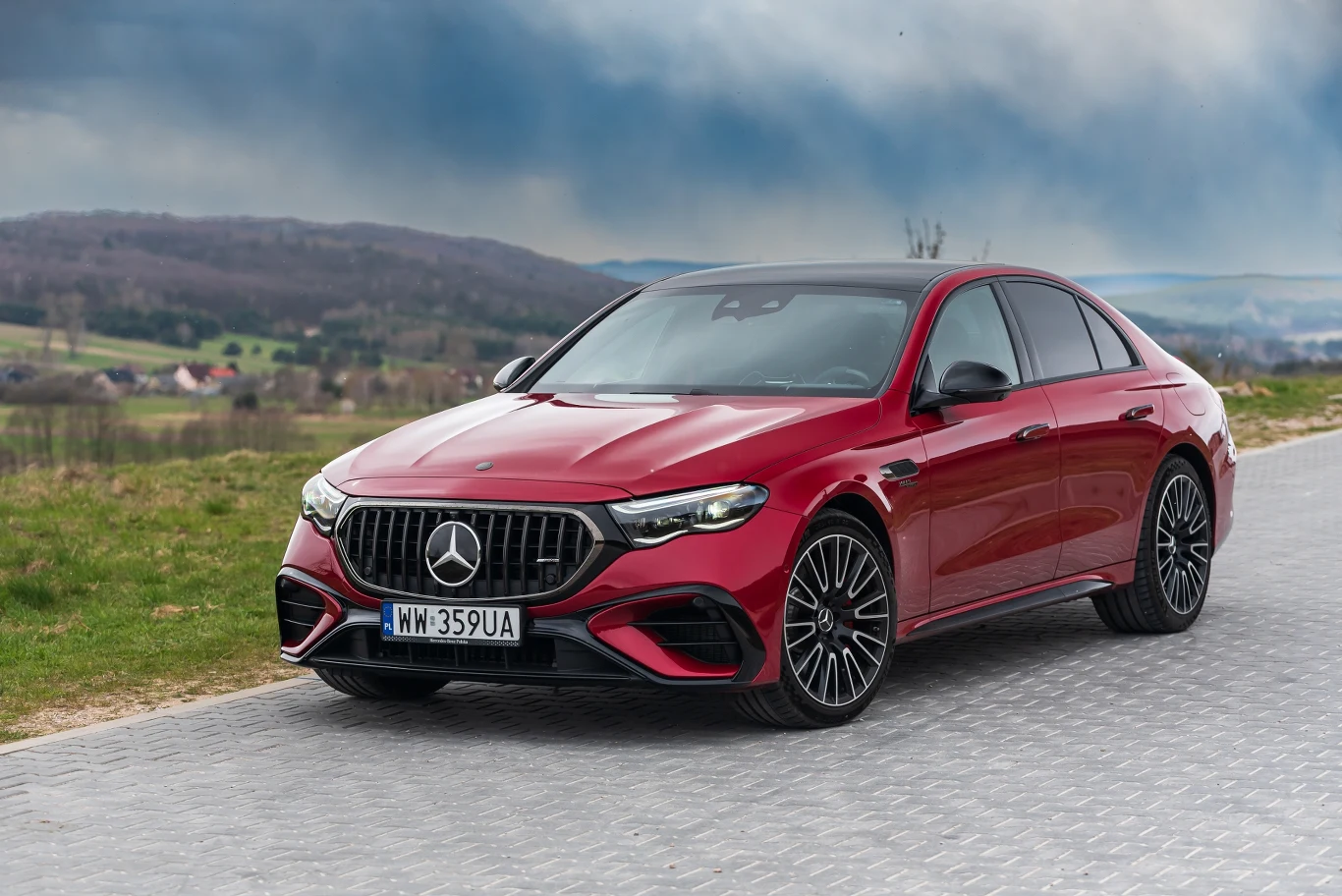 Czerwony Mercedes-Benz klasy E stojący na brukowanej drodze, otoczony zielonym krajobrazem z widocznymi wzgórzami i pochmurnym niebem w tle.