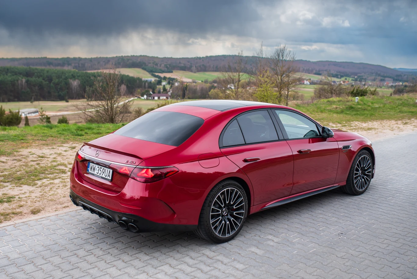 Czerwony luksusowy sedan z przyciemnianymi szybami stoi na brukowanej drodze na tle malowniczego krajobrazu z polami, lasem i wzgórzami pod pochmurnym niebem.