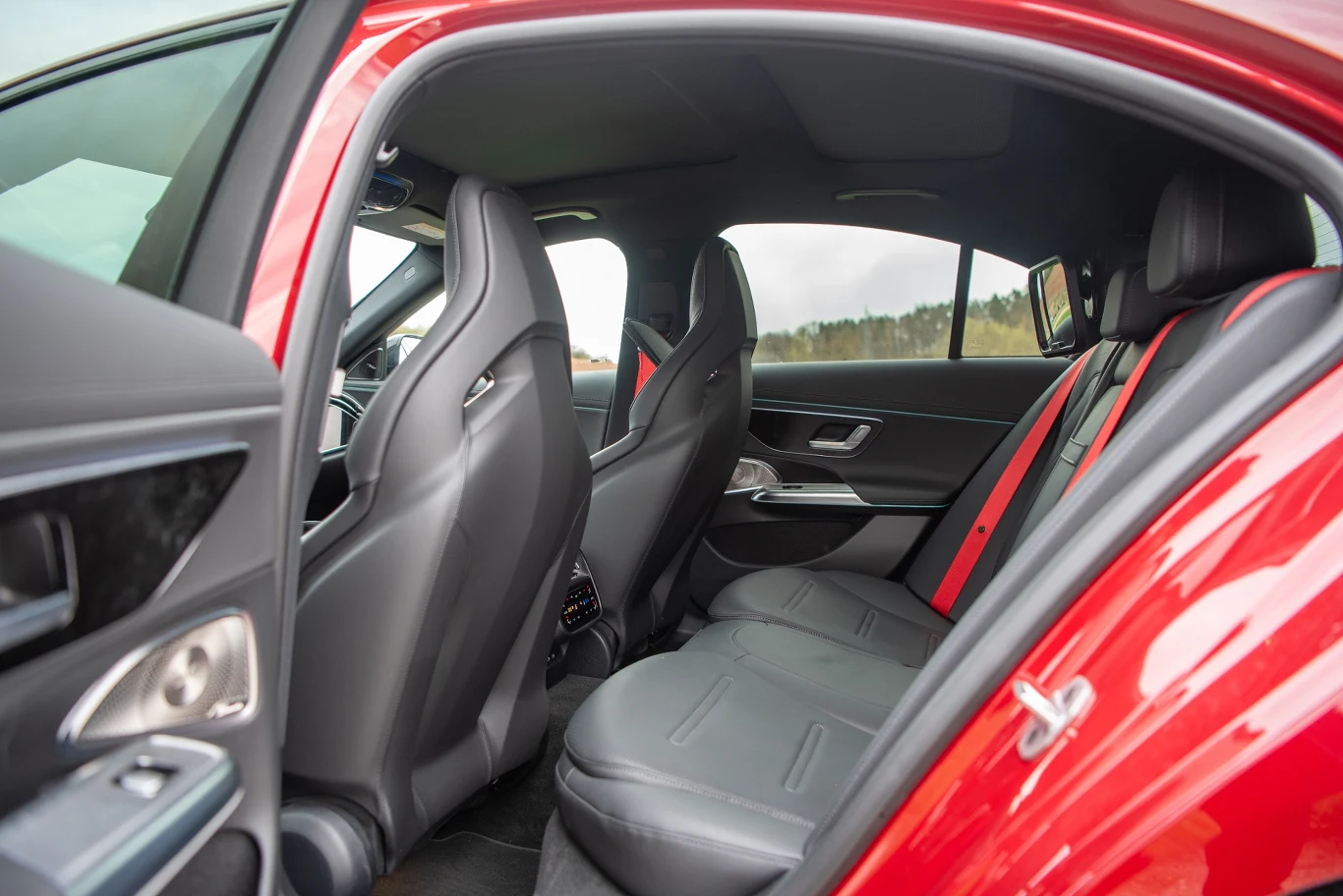 Obszerne wnętrze samochodu osobowego z czerwonymi pasami bezpieczeństwa, skórzanymi czarnymi fotelami oraz nowoczesnym wykończeniem drzwi i paneli bocznych. Ujęcie z tylnej części samochodu, przez otwarte drzwi.