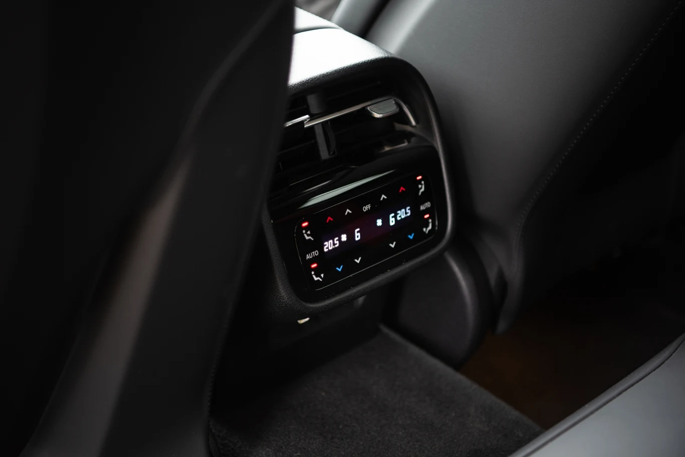 Panel sterowania klimatyzacją zamontowany na tylnej konsoli samochodu, z cyfrowym wyświetlaczem oraz przyciskami do regulacji temperatury i nawiewu, otoczony skórzanymi fotelami.