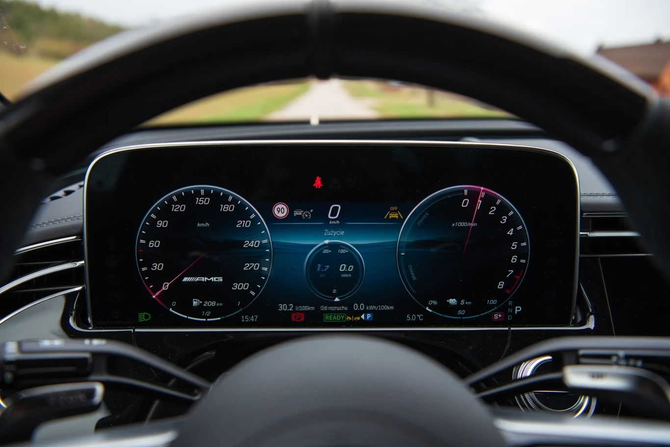 Cyfrowy panel wskaźników luksusowego samochodu pokazuje aktualną prędkość, poziom paliwa, informacje dotyczące jazdy oraz ustawień pojazdu, wraz z wyraźnie widoczną kierownicą i drogą w tle.