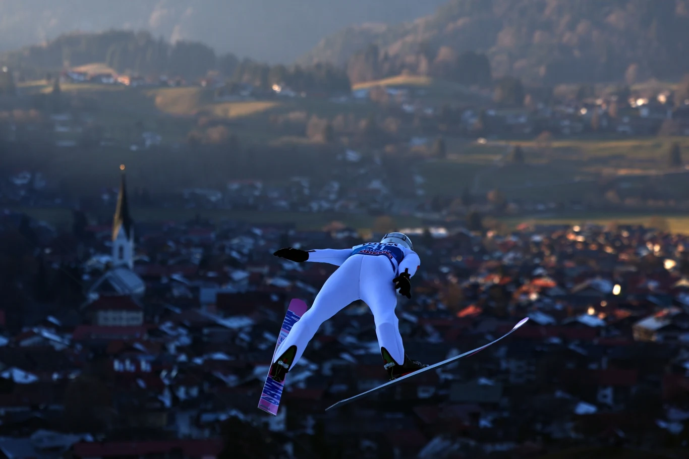 Skoczek narciarski wykonujący skok na tle zabudowań górskiego miasteczka, w oddali widać rozległy krajobraz, kościół oraz lekko zamglone wzgórza.