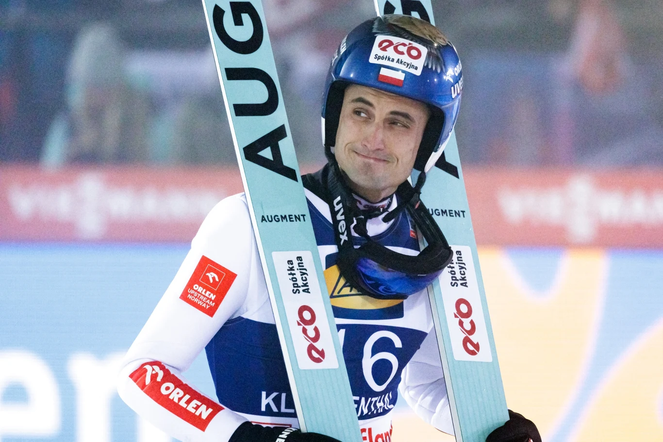 Skoczek narciarski uśmiechający się w stroju sportowym, trzyma narty i stoi na tle rozmytej publiczności oraz reklam bandowych.