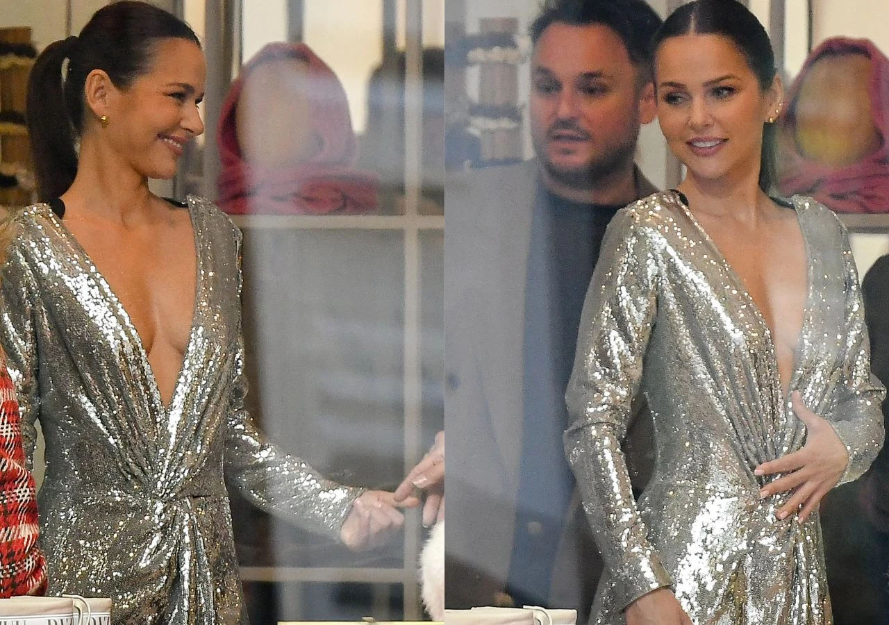 Kobieta w połyskującej srebrnej sukni z głębokim dekoltem, stojąca w towarzystwie mężczyzny w eleganckim stroju. Para znajduje się w jasnym pomieszczeniu z widocznymi detalami wnętrza.