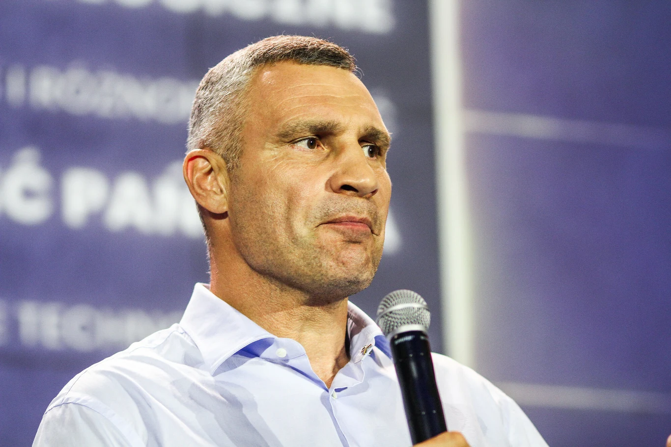 Mężczyzna w jasnej koszuli trzyma mikrofon i patrzy w bok, w tle rozmazane napisy na niebieskim tle.