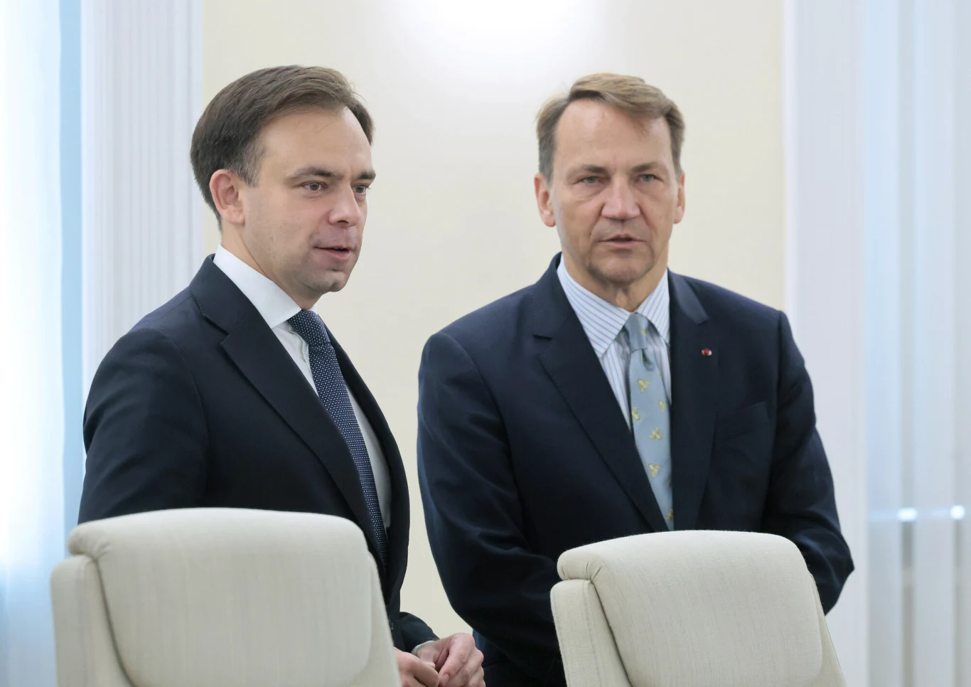 Bułgaria przyjmuje euro. Sikorski chwali korzyści, Domański mówi "nie" (zdj. ilustracyjne) Dwóch mężczyzn w garniturach stoi obok siebie w jasnym, formalnym pomieszczeniu. Obaj skierowani są lekko bokiem, rozmawiają ze sobą, na pierwszym planie widoczne są jasne oparcia krzeseł.