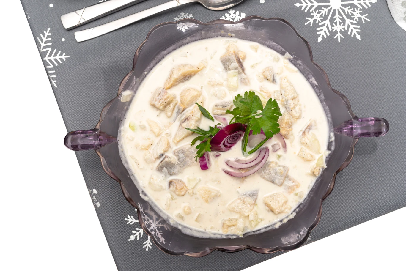 Kawałki śledzia w śmietanowym sosie z dodatkiem czerwonej cebuli oraz natki pietruszki, podane w przezroczystej miseczce na szarej serwetce ze śnieżynkami.