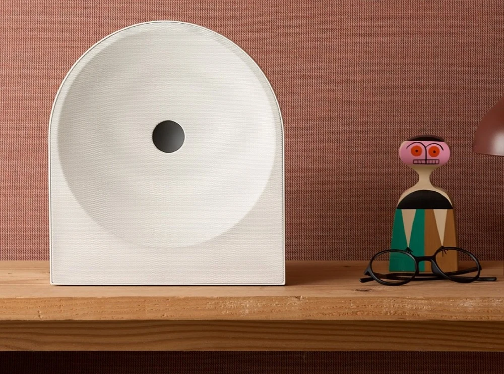 Music Studio to nowa seria głośników, które Samsung pokaże na CES 2026. Biała, geometryczna rzeźba z okrągłym otworem ustawiona na drewnianej półce przy ścianie w kolorze ceglastym, obok drewniana figurka o stylizowanych, kolorowych kształtach, poniżej okulary oraz lampa widoczna z prawej strony kadru.