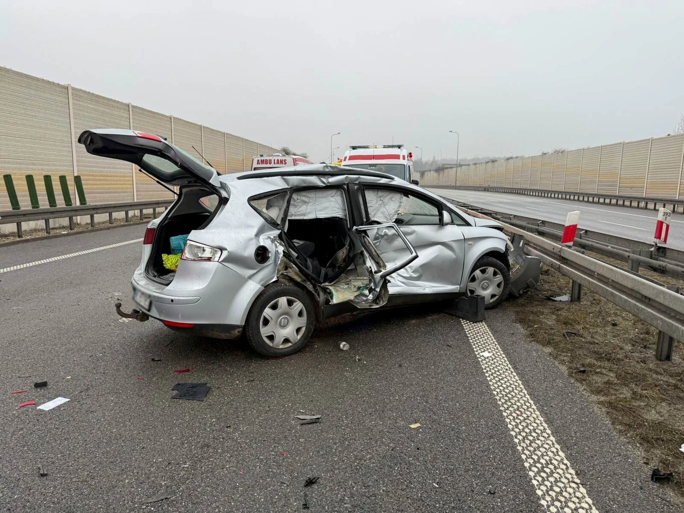 Rozbity samochód osobowy marki taxi stoi na pasie autostrady przy barierkach ochronnych, z poważnymi uszkodzeniami po stronie kierowcy i otwartym bagażnikiem. W tle znajduje się ambulans, na drodze widoczne są ślady zdarzenia oraz elementy pojazdu.