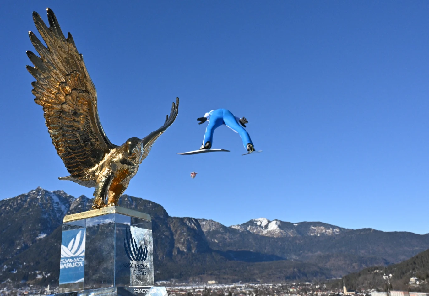 Złoty orzeł jako trofeum w konkursie skoków narciarskich na pierwszym planie, wysoko lecący skoczek narciarski ubrany w niebieski kombinezon na tle gór oraz błękitnego nieba w tle.