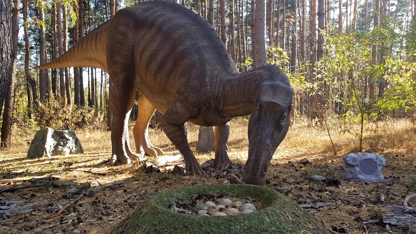 Dinozaur stojący w lesie, pochylony nad gniazdem z jajami, otoczony drzewami i suchą ściółką leśną.