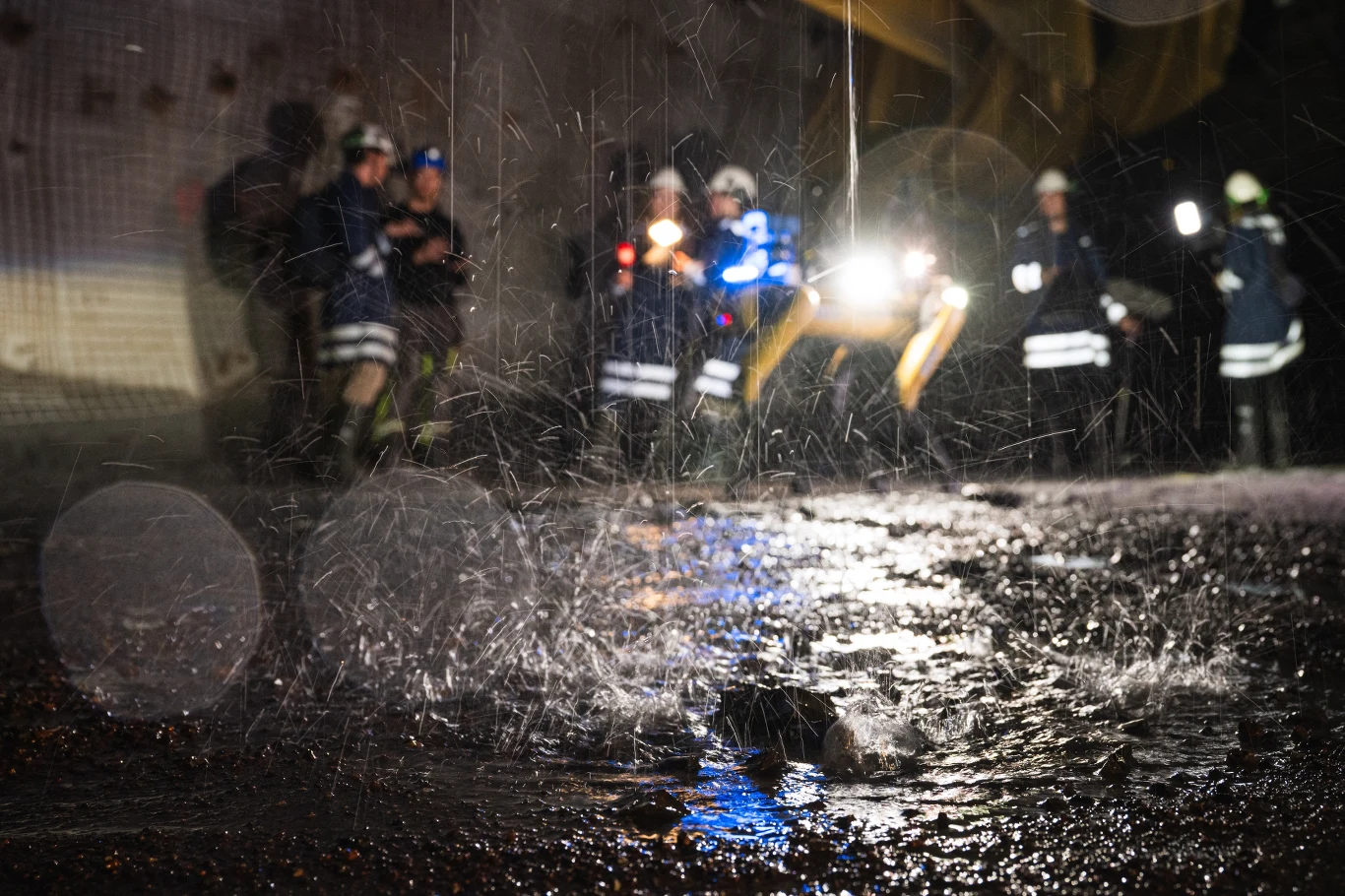 Strażacy w odblaskowych strojach stojący w tle podczas intensywnych opadów deszczu, na pierwszym planie widoczna ulewa i spływająca po ziemi woda, w tle światła pojazdów ratowniczych.
