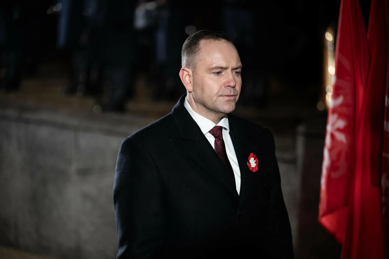 Mężczyzna w eleganckim ciemnym płaszczu i bordowym krawacie z przypiętym kokardą narodową, stojący obok czerwonej flagi, w tle rozmyte sylwetki ludzi oraz kamienne schody.