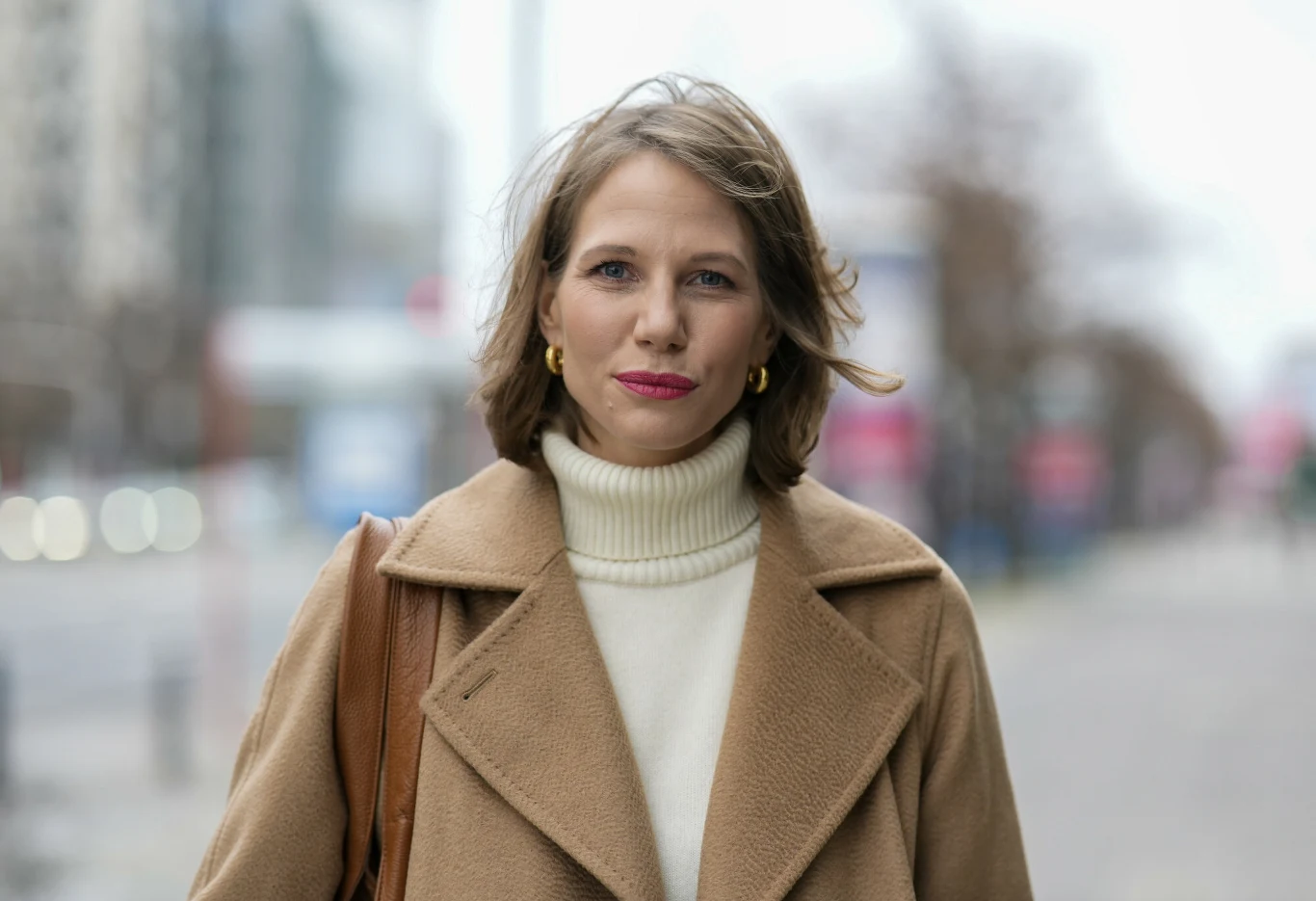 Kobieta z rudobrązowymi włosami ubrana w beżowy płaszcz i jasny golf, stojąca na tle miejskiej ulicy. Wyraz twarzy spokojny, delikatny uśmiech, złote kolczyki w uszach. W tle rozmazana zabudowa miejska.