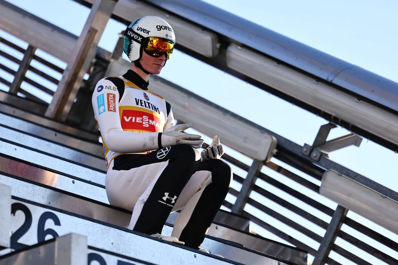 Skoczek narciarski w kombinezonie startowym i kasku siedzący na belce startowej skoczni, skupiony przed oddaniem skoku, w tle konstrukcja skoczni.