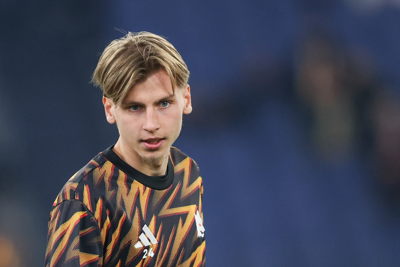 Młody mężczyzna z jasnymi włosami ubrany w sportową bluzę z geometrycznym wzorem i logo Adidasa na tle rozmytego, ciemnoniebieskiego tła.