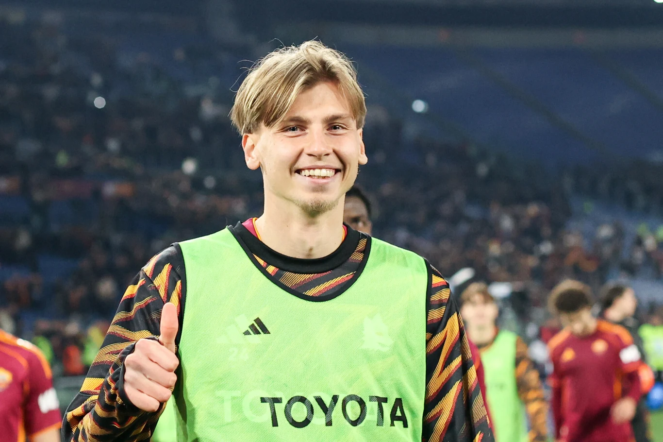 Młody mężczyzna w sportowej bluzie i zielonej kamizelce z logo Toyota uśmiecha się i pokazuje kciuk w górę. W tle widać rozmazany stadion z kibicami oraz innych zawodników w strojach sportowych.