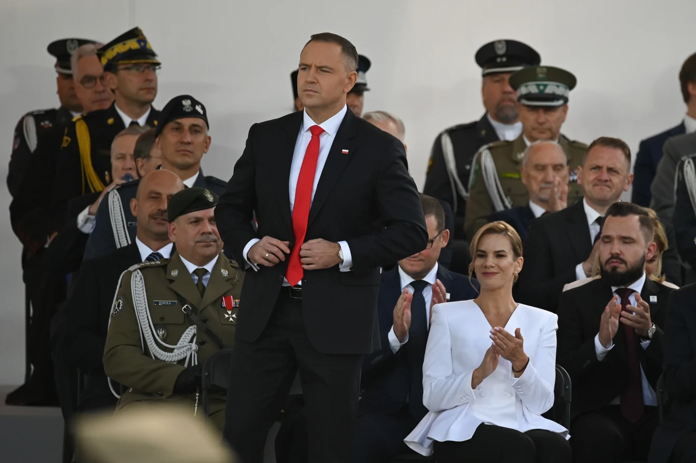 Prezydent stoi na tle innych uczestników uroczystości, ubrany w granatowy garnitur i czerwoną krawatę, obok klaskająca kobieta w białej marynarce. W tle widoczni są oficerowie wojska i służb mundurowych oraz inni oficjele siedzący w kilku rzędach.