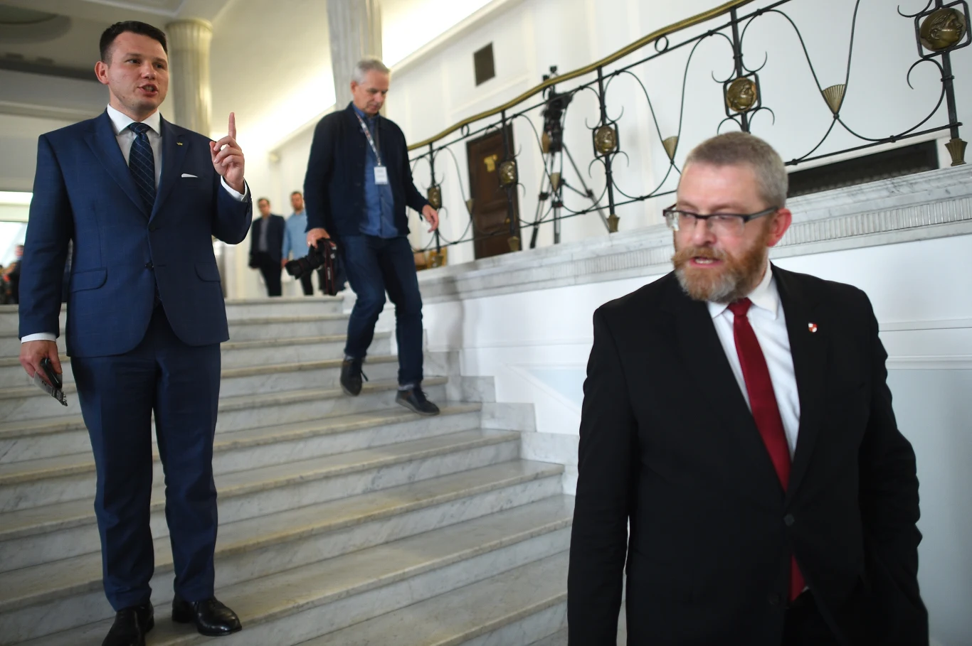 Trzech mężczyzn w garniturach na marmurowych schodach w budynku o reprezentacyjnym wnętrzu, jeden z nich stoi i gestykuluje, drugi schodzi w dół bliżej kamery, a trzeci idzie w tle z aparatem fotograficznym.