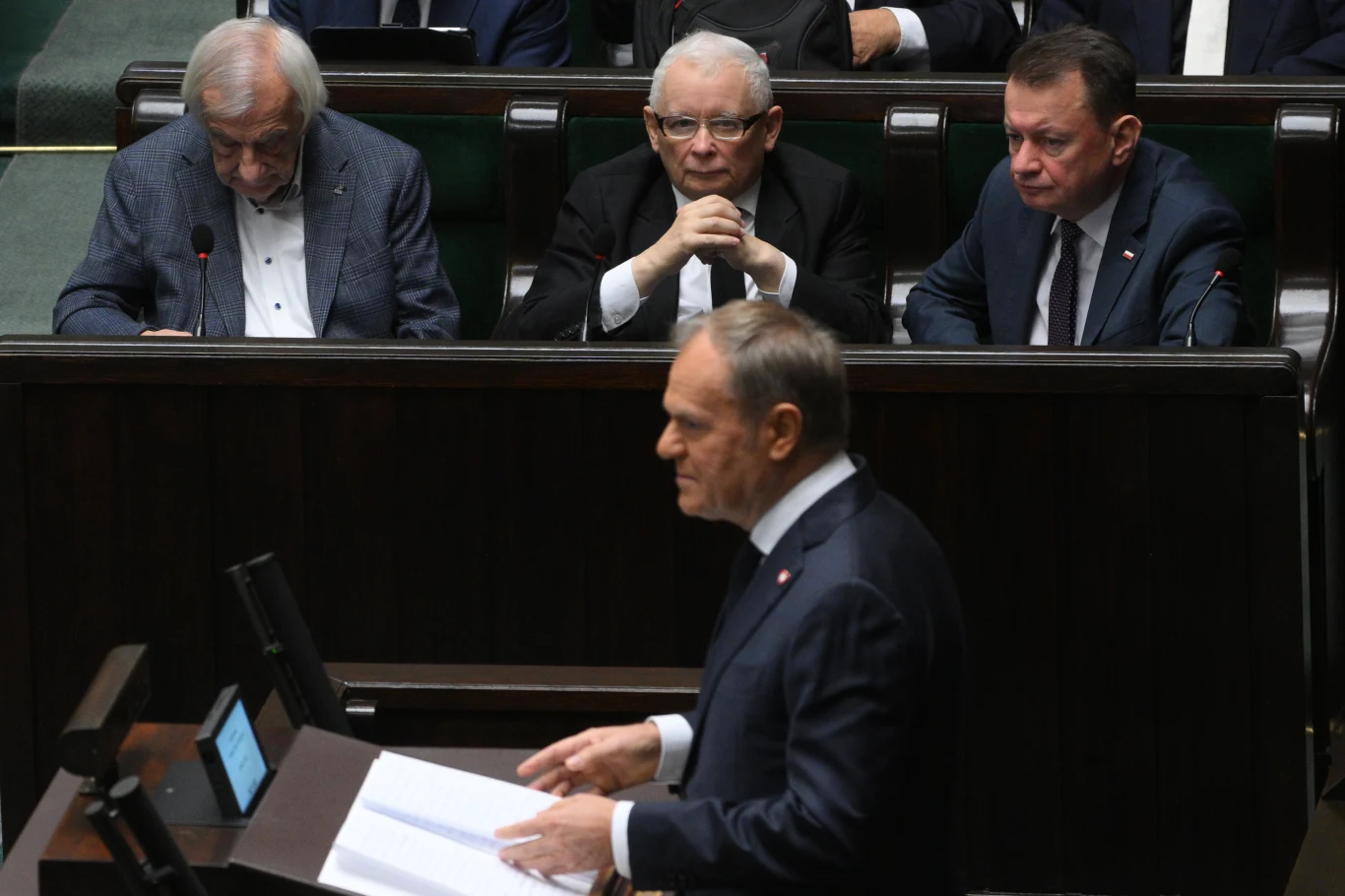 Premier przemawia przy mównicy podczas obrad w parlamencie, na pierwszym planie znajduje się mężczyzna w garniturze z dokumentami, w tle trzech polityków uważnie słucha wystąpienia.