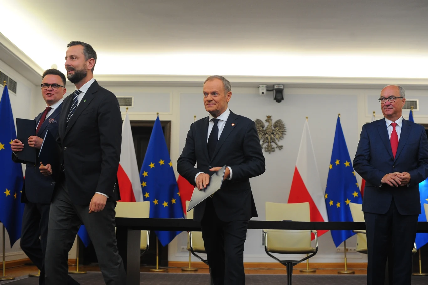 Czterech mężczyzn w garniturach w sali konferencyjnej, w tle flagi Polski i Unii Europejskiej oraz godło Polski, atmosfera oficjalna, przedstawiciele władz biorą udział w uroczystości.