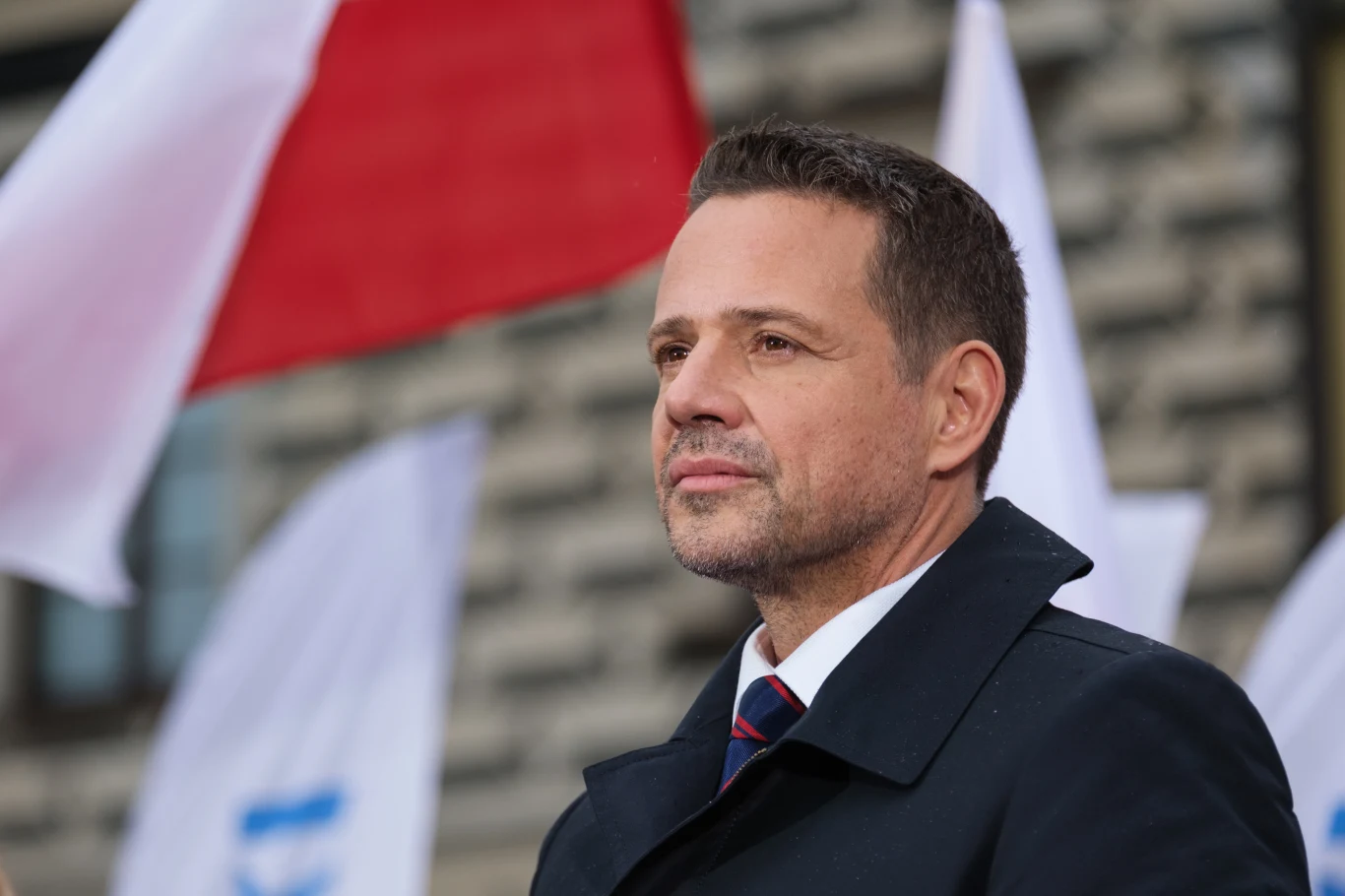 Mężczyzna w garniturze i płaszczu patrzy w dal z wyrazem skupienia. W tle widoczne są biało-czerwone i białe flagi, rozmyty budynek oraz jasne tło.