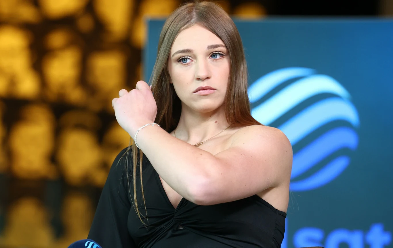 Młoda kobieta o długich, jasnych włosach poprawia fryzurę. Ubrana w czarną sukienkę, stoi na tle niebieskiego logo przypominającego fale na rozmytym tle.