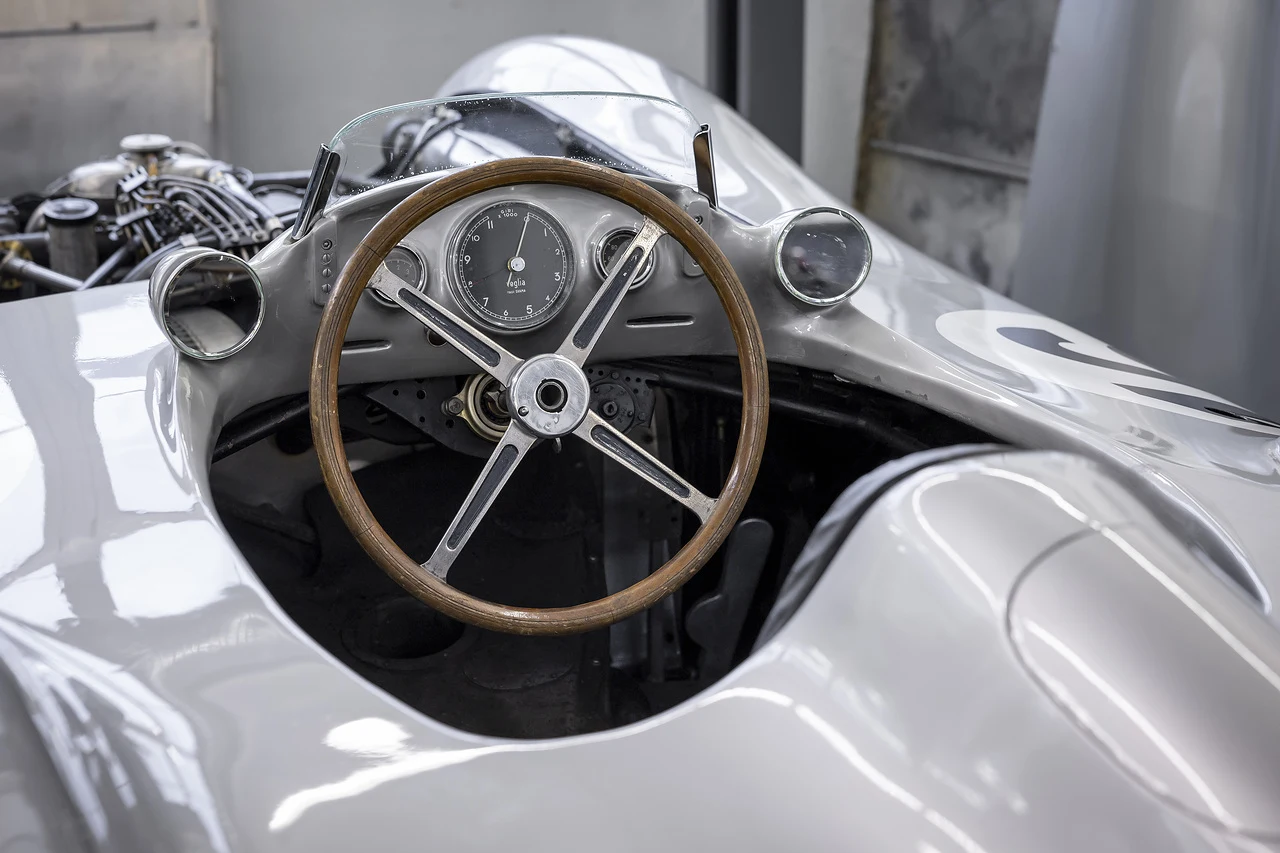 Kokpit klasycznego samochodu wyścigowego z drewnianą kierownicą, analogowym prędkościomierzem oraz widocznością części silnika po lewej stronie.