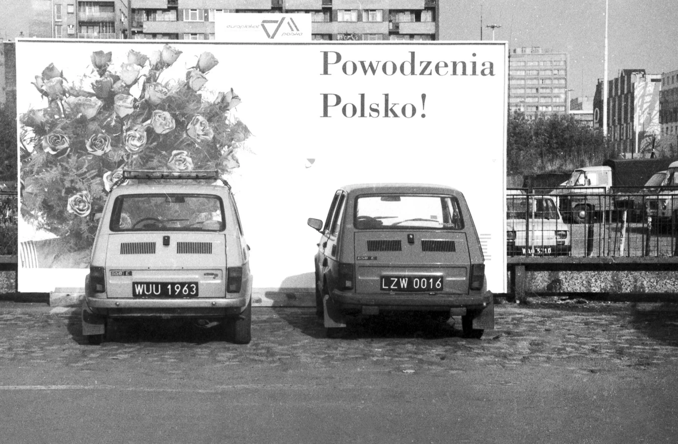 Dwa samochody osobowe zaparkowane przed dużym billboardem z napisem Powodzenia Polsko i grafiką bukietu róż. W tle widoczne fragmenty budynków mieszkalnych oraz miejski krajobraz.